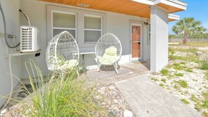 Una casa moderna con un porche cubierto, dos sillas blancas con forma de huevo y un equipo de aire acondicionado blanco.