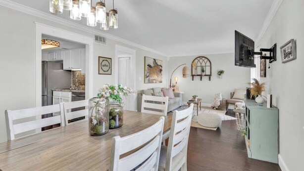 Een eethoek met een houten tafel, witte stoelen en een kroonluchter. In de achtergrond is een keuken zichtbaar met witte kasten en een koelkast.