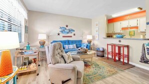 Una sala de estar con un sofá azul, un sillón color beige, una mesa de centro y una cocina con taburetes rojos.