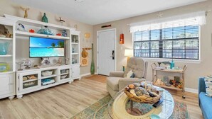 Una sala de estar con televisor, mueble blanco para entretenimiento y ventana con persianas.