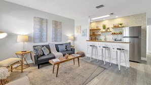 Ein modernes Wohnzimmer mit grauem Sofa, einem Holztisch und einer Küchenzeile mit Edelstahl-Kühlschrank, Barhocker und einer Arbeitsplatte mit Regalen.