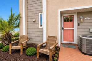 Un bâtiment avec deux chaises en bois à l’extérieur, une porte rouge et un patio agrémenté d’un petit jardin.