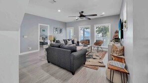 A spacious living room with a grey sofa, a ceiling fan, and a large window providing natural light. - Large 3BR Family Destin Beach House w Beach Access (Destin)