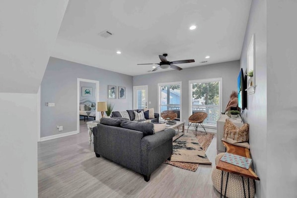 A spacious living room with a grey sofa, a ceiling fan, and a large window providing natural light.