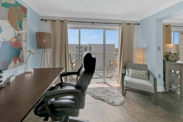 A room with a dining table, a black office chair, a sofa, and a balcony with a view.