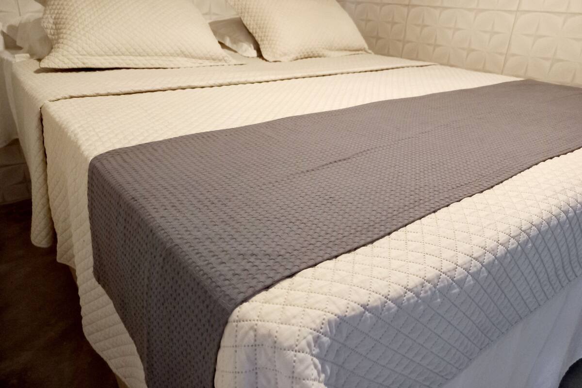 Ein Doppelbett mit weißgrauer gesteppter Bettdecke, einem weißen Kopfteil und einer Wand mit Muster.