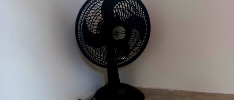A black standing fan with a circular grill and a base, positioned against a white wall on a concrete floor.