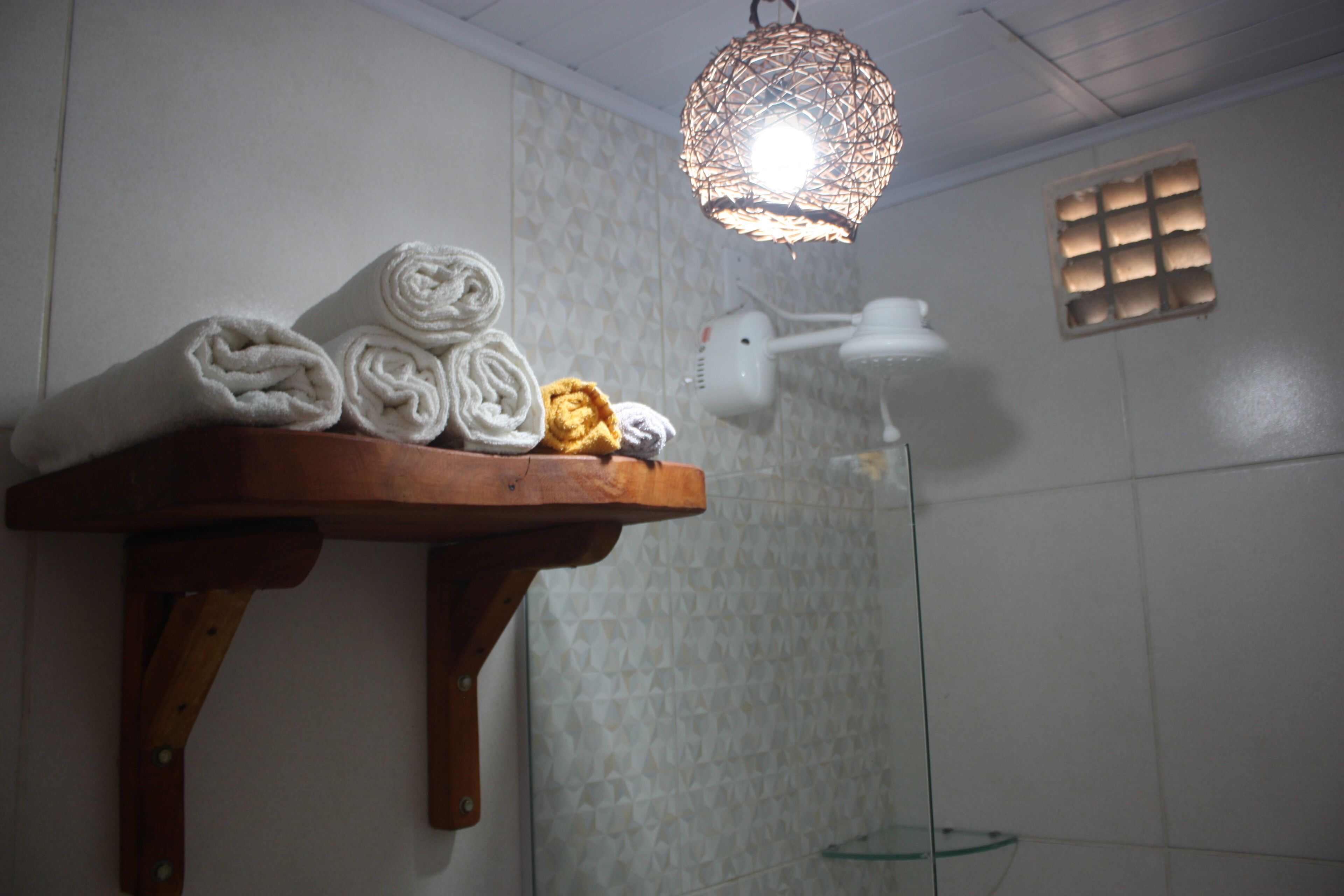 Une étagère en bois avec des serviettes enroulées et un sèche-cheveux, dans une salle de bain aux carreaux décorés et dotée d’un luminaire suspendu.