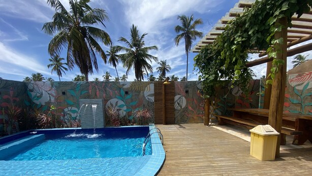 A pool area with a wooden deck, a mural with tropical leaf patterns, and palm trees in the background. - Chalé Palhano Beach Milagres (São Miguel dos Milagres)