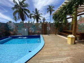 A pool area with a wooden deck, a mural with tropical leaf patterns, and palm trees in the background. - Chalé Palhano Beach Milagres (São Miguel dos Milagres)