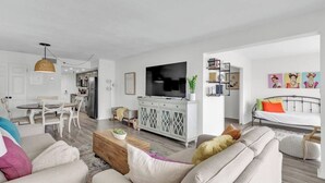 A modern living room with a sofa, coffee table, and a TV. There is a dining area with a table and chairs, and a kitchen in the background.