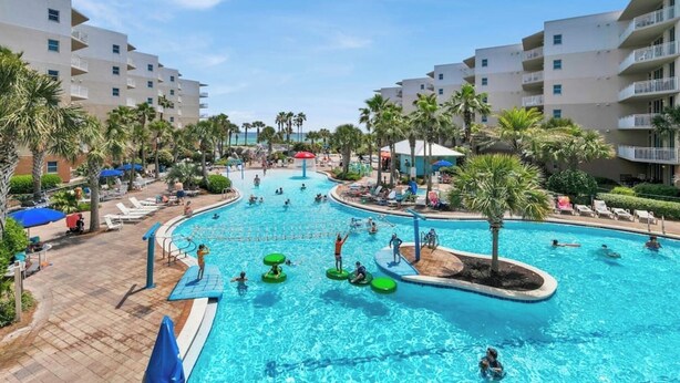 A large outdoor swimming pool surrounded by palm trees and lounge chairs, with multiple levels of buildings in the background.