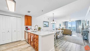 A modern kitchen with white cabinets, a central island, and wooden accents. A dining area with a table and chairs is visible, and a living room with a sofa and a view of the city is in the background.