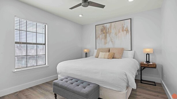 Une chambre à coucher avec un lit, deux tables de chevet avec des lampes, un ventilateur de plafond, une fenêtre avec des stores et un grand tableau abstrait accroché au mur.