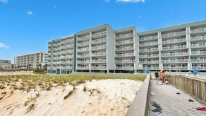 Un hotel frente a la playa con varios balcones, un paseo marítimo de madera y una playa arenosa en primer plano.