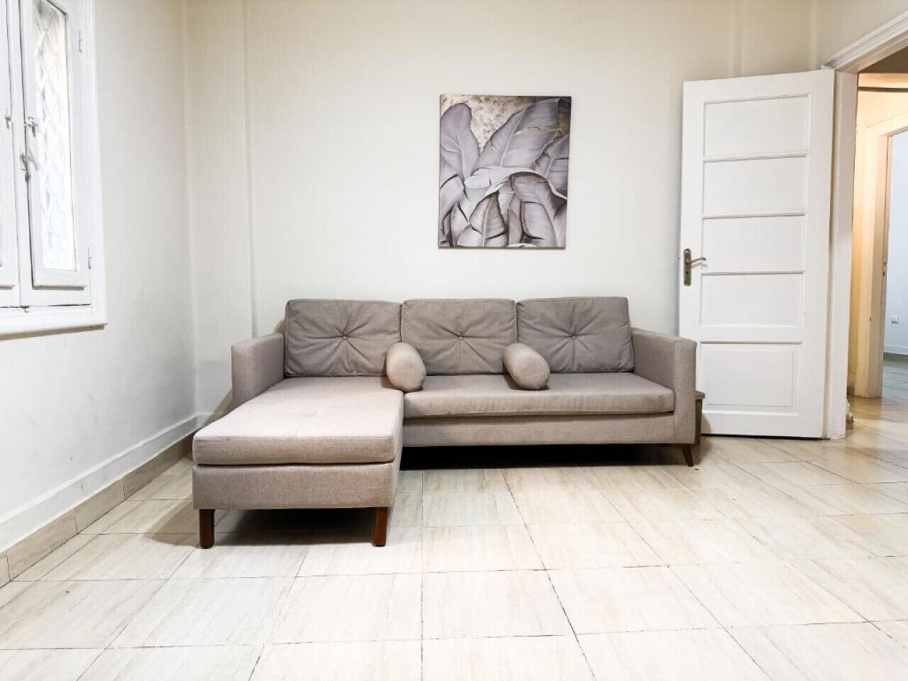 Un sofá de tonos neutros con un reposapiés a juego, una obra de arte abstracta colgada en la pared y una puerta blanca en una habitación con suelo de baldosas de colores claros.