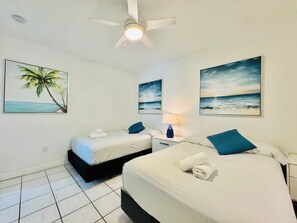 Ein Hotelzimmer mit zwei Betten, einem Deckenventilator und Kunstwerken, die Strandmotive zeigen.