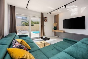 Un salon moderne avec un canapé vert, un téléviseur à écran plat et une porte-fenêtre coulissante donnant sur une piscine.