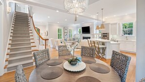A spacious living area with a wooden staircase, a chandelier, wicker furniture, and a dining table with placemats.