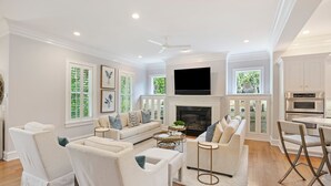 A spacious living room with a fireplace, a flat-screen TV, and a dining area with a kitchen in the background.