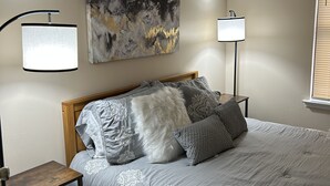 Quarto com cama, duas mesas de cabeceira, um quadro abstrato na parede e uma janela com persianas.