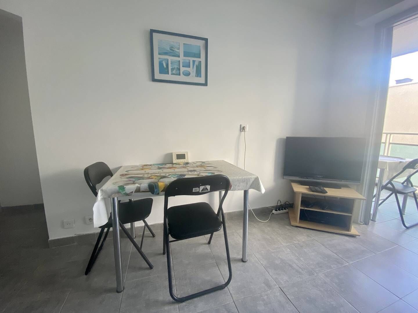 En liten matplats med ett bord och stolar, en inramad tavla på väggen och en tv på ett stativ.