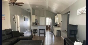 Una sala de estar con un sofá, un televisor y un ventilador de techo. Hay una zona de cocina con armarios y un microondas.