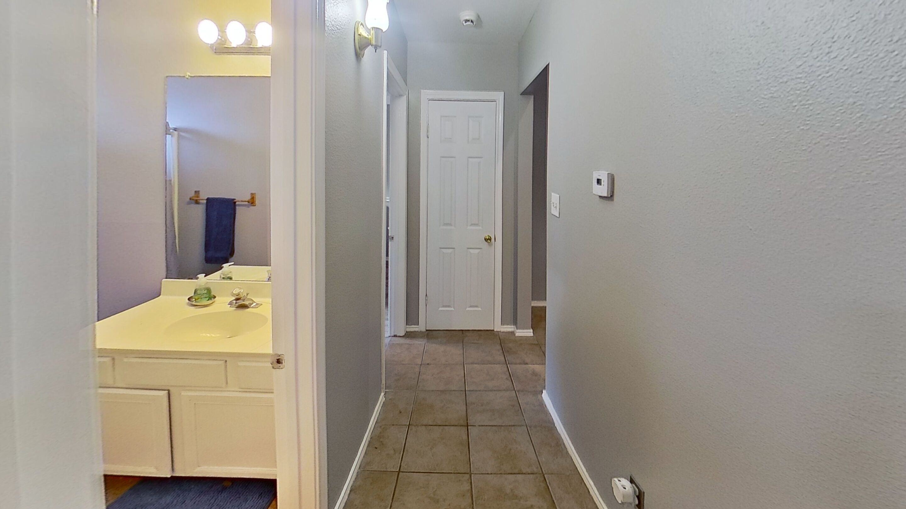 A narrow hallway with a bathroom on the left, featuring a sink and mirror. A white door is visible at the end of the hallway.