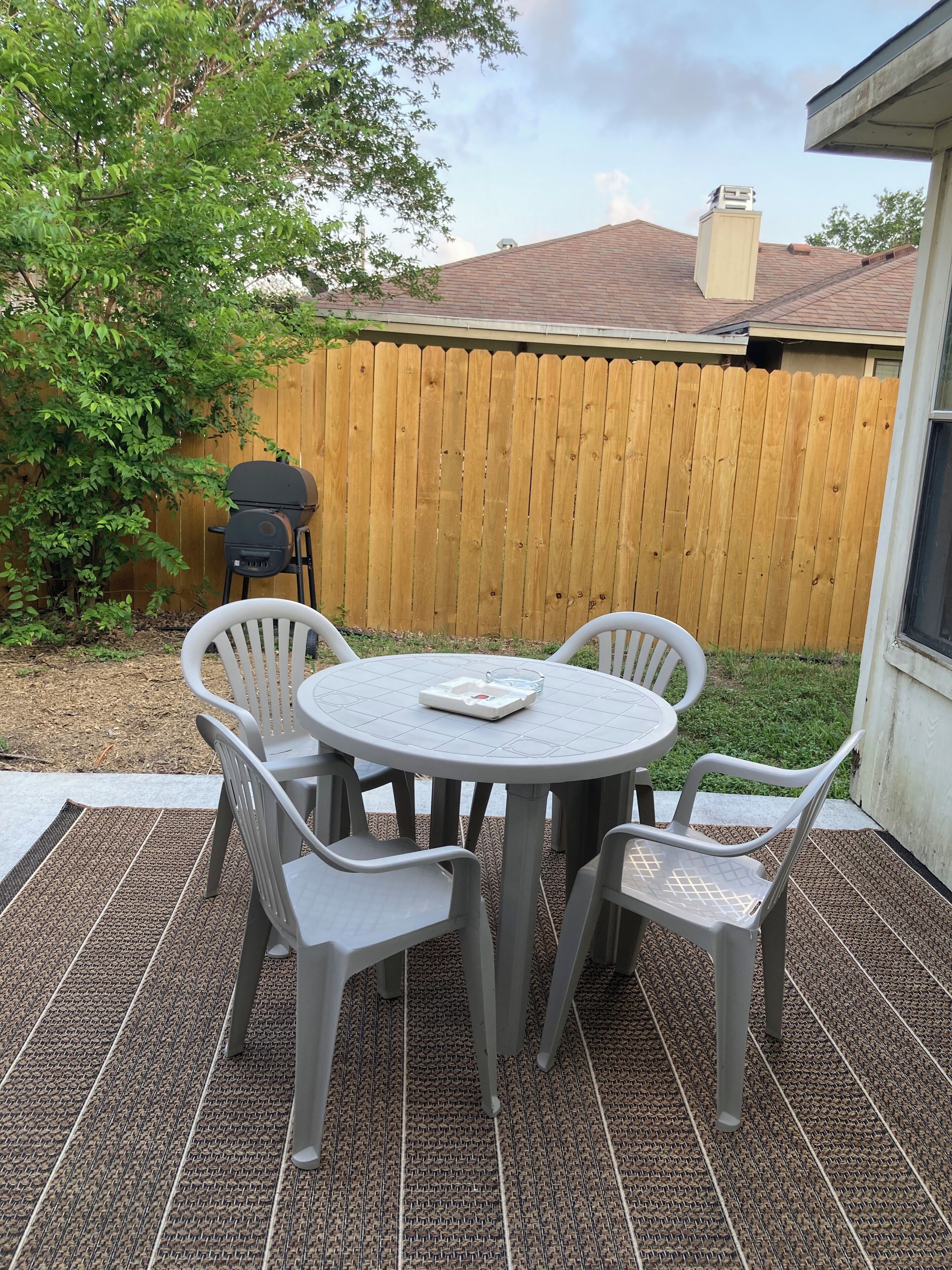Un patio con una mesa redonda y cuatro sillas, una cerca de madera y una parrilla al fondo.