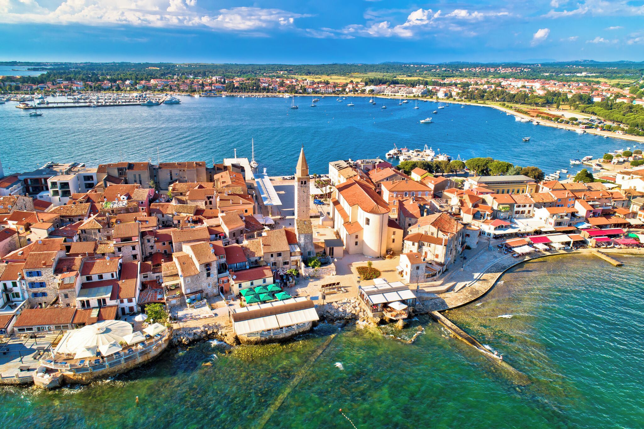 Vista aérea de un pueblo costero con edificios históricos, una iglesia destacada y un puerto deportivo con barcos.