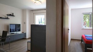 Una habitación moderna con televisión de pantalla plana, un escritorio y una silla. Hay una cama con colcha roja y azul. La habitación tiene un ventanal grande y una puerta que da a otra estancia.