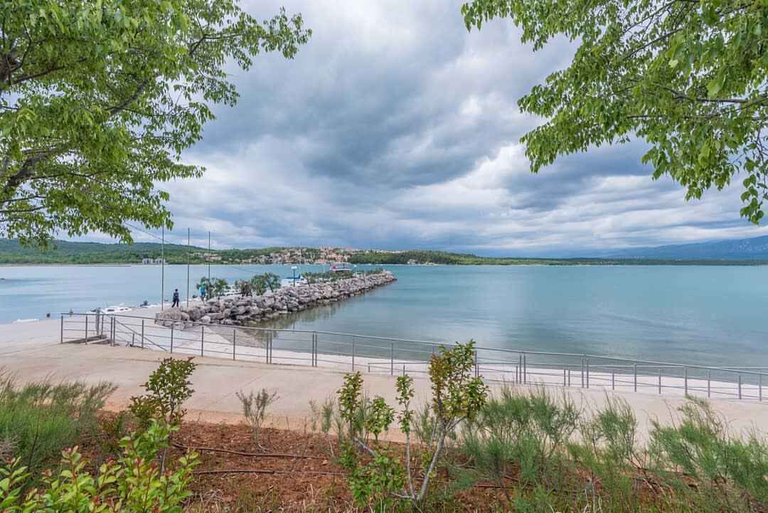 A coastal walkway with a rocky breakwater, a clear view of the sea, and a distant town.