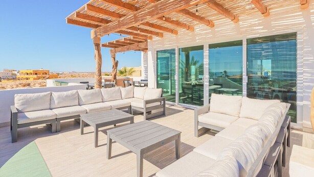 An outdoor seating area with a wooden pergola, white sofas, and a glass wall overlooking the beach.
