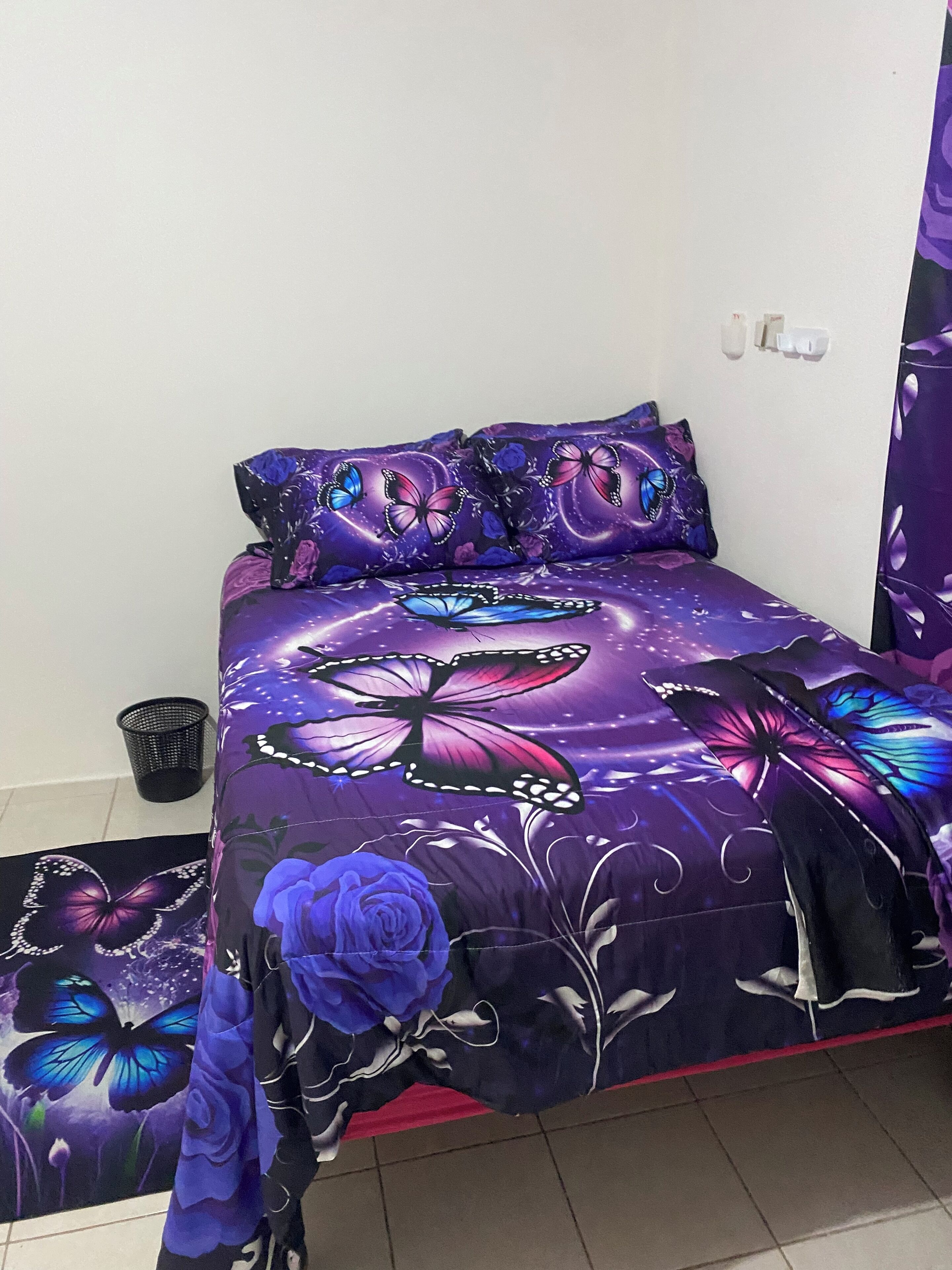 A bed with a purple and blue butterfly and floral design, a black trash bin, and a red rug.