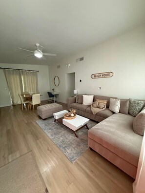 Un salon avec un canapé, une table basse et un ventilateur de plafond. Une pancarte sur le mur indique : « MAISON DE PLAGE ».