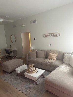 A modern living room with a sofa, ottoman, coffee table, and a desk with a chair. A sign on the wall reads 'BEACH HOUSE.'