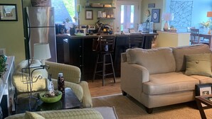 Un salon confortable avec un canapé et des fauteuils, un coin cuisine équipé d’un réfrigérateur et d’un plan de travail, et un coin repas avec une table et des chaises.