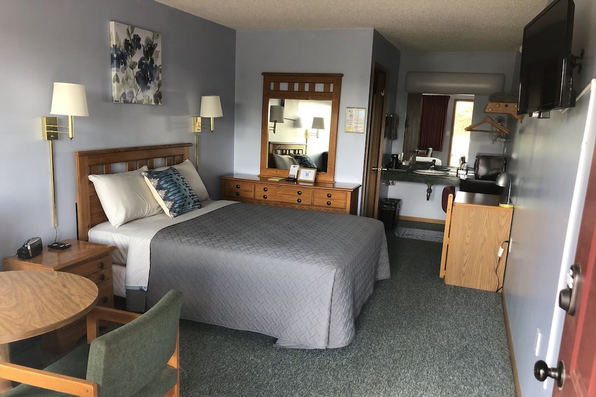 A compact hotel room with a bed, bedside tables, a desk, a chair, a mirror, a sink, and a television.
