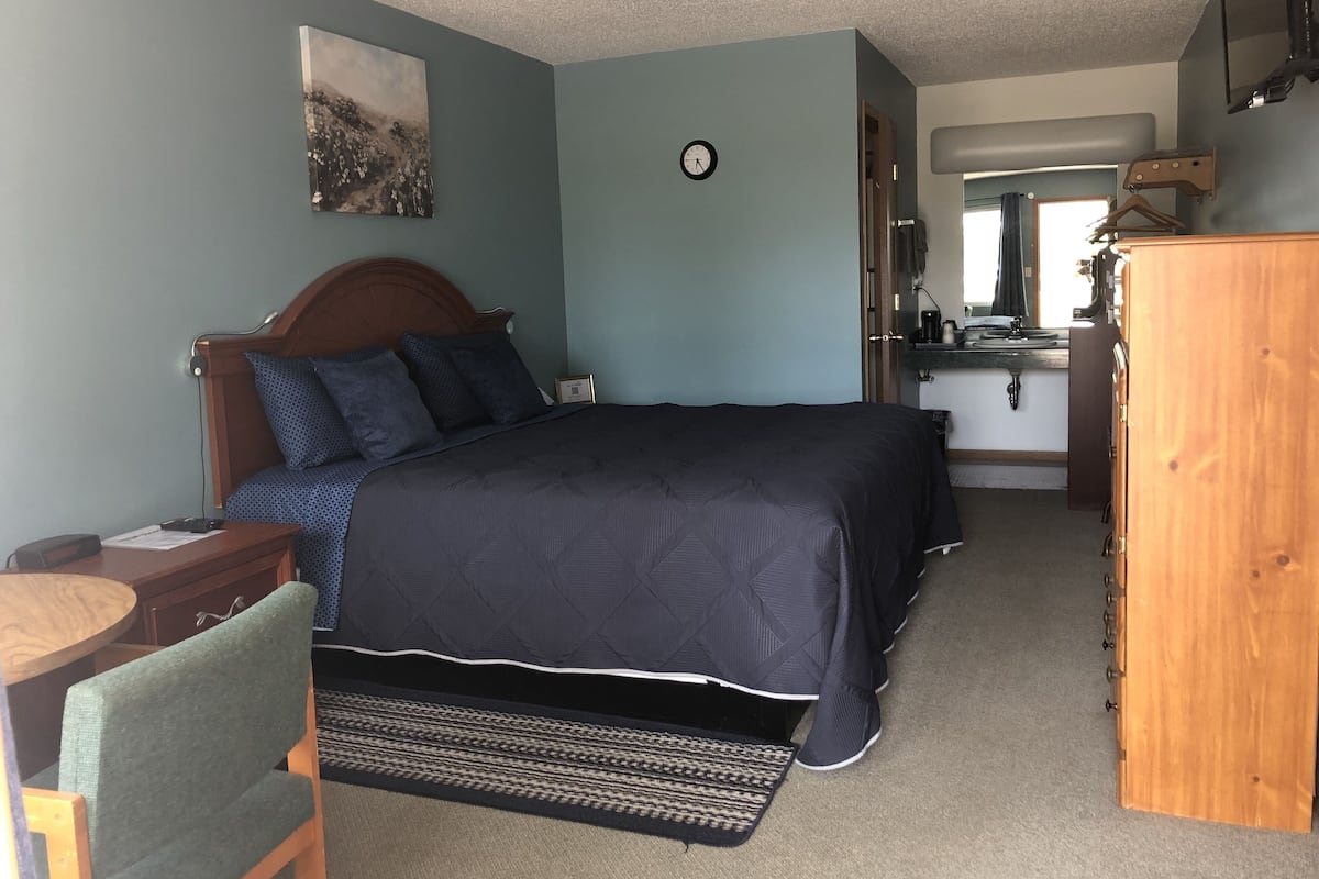 A bedroom with a bed, a desk, a chair, a dresser, a clock, and a painting on the wall.