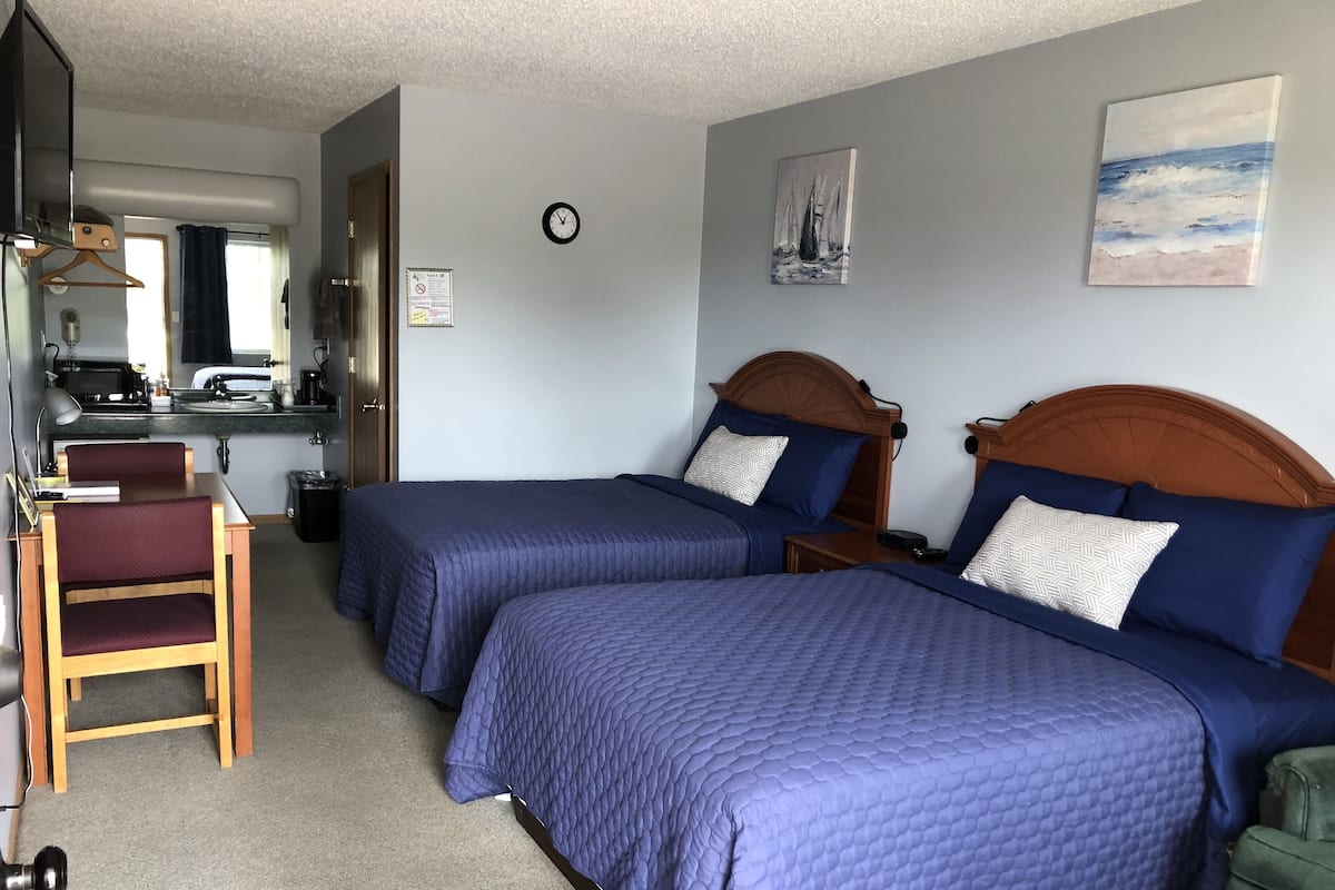 A hotel room with two beds, a desk, a chair, a clock, and a painting on the wall.