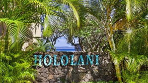 Un mur de pierre sur lequel est inscrit en grandes lettres le nom «  HOLOLANI », entouré d’une végétation luxuriante et offrant une vue sur l’océan.
