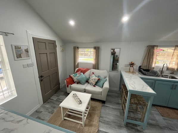 A compact living space with a sofa, a small table, a kitchen area, and a window with curtains.