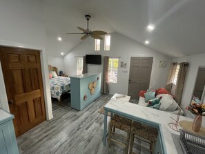 A cozy, well-lit room with a bed, a small kitchenette, a dining area, and a ceiling fan.
