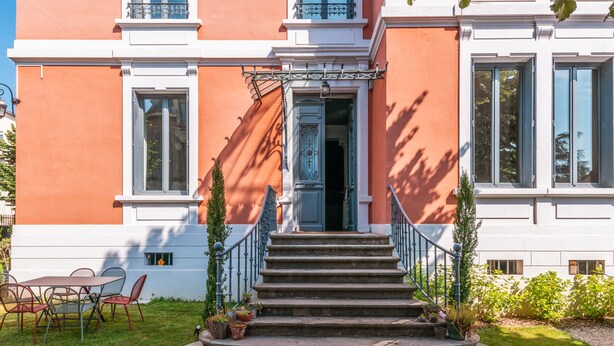Un edificio con fachada roja, detalles blancos y una puerta azul. Hay escalones que conducen a la entrada y un pequeño jardín con plantas en macetas.