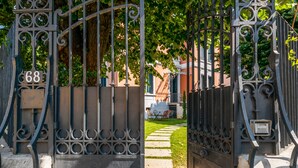 Una entrada con portón, una placa con el número 68, un camino pavimentado y un edificio al fondo.