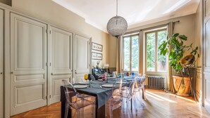 Un comedor con suelo de madera en espiga, una mesa con sillas transparentes, una araña y un ventanal con vistas a zonas verdes.