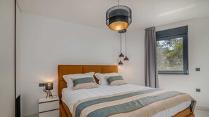 A modern bedroom with a wooden headboard, a large bed with striped bedding, bedside lamps, and a view of greenery through a window. - Apartment AK lux with private pool (Njivice)