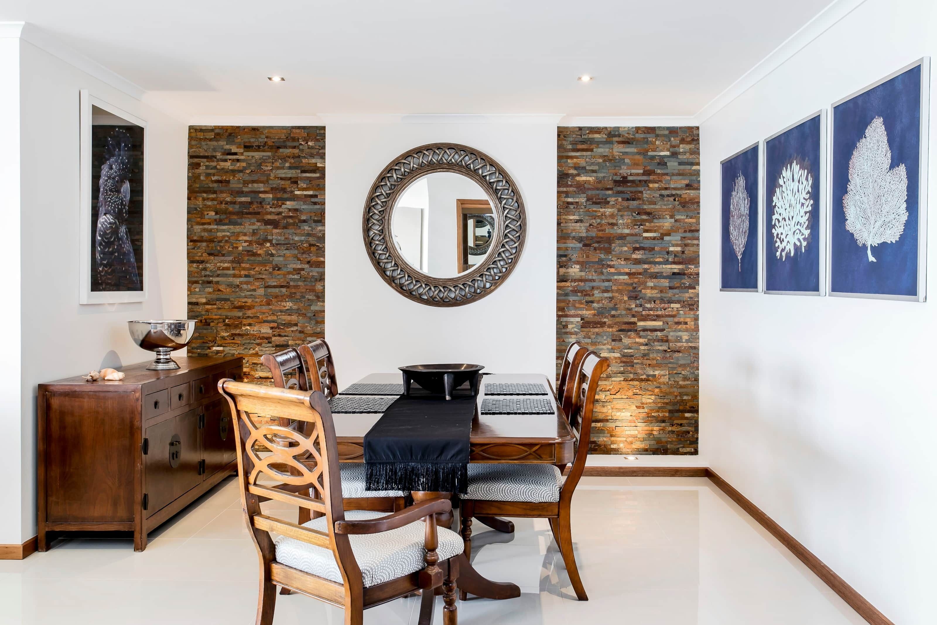 A dining area with a wooden table, chairs, a central mirror, and a decorative wall with a brick pattern.