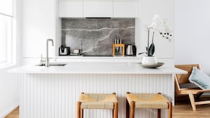 Une cuisine moderne avec un plan de travail blanc, un pare-feu en marbre et un évier encastré. On y trouve deux tabourets en osier et une chaise en bois avec un coussin.