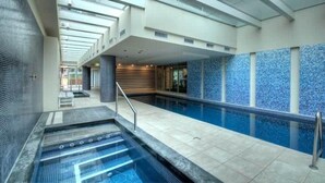 Una piscina interior moderna con techo de cristal, una zona de hidromasaje separada y una pared de azulejos con un diseño de mosaico azul.